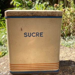 Op de afbeelding: Een vintage witte metalen suikerpot met blauwe en oranje strepen en het woord "SUCRE" in het blauw.