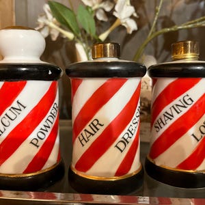 Könnte beinhalten: Set mit drei Apothekergläsern aus einem Vintage-Friseursalon. Jedes Glas ist weiß mit roten und weißen diagonalen Streifen, schwarzen Akzenten und goldenen Verzierungen. Die Gläser sind mit "Talcum Powder", "Hair Dressing" und "Shaving Lotion" beschriftet.