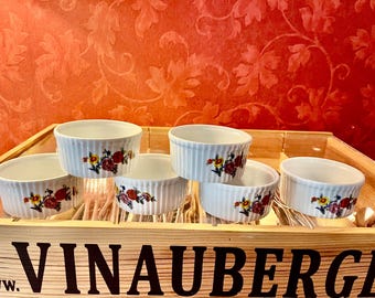 May include: Six white ceramic ramekins with floral designs in red, yellow, and green. The ramekins are arranged in two rows of three on a wooden surface with the text "VINAUBERG" printed on the surface.