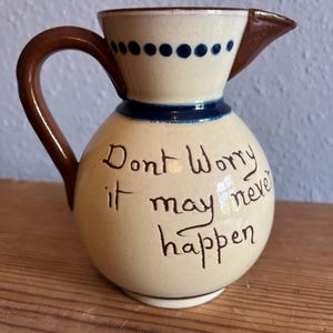 May include: A cream-coloured ceramic pitcher with a brown handle and rim. The pitcher features a dark blue dotted pattern around the neck and the hand-painted brown text "Dont Worry it may never happen" on the front of the rounded body.