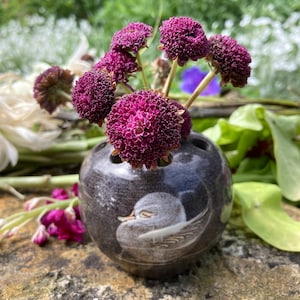 May include: A small, dark gray ceramic vase with a white duck design. The vase is filled with a bouquet of purple flowers.