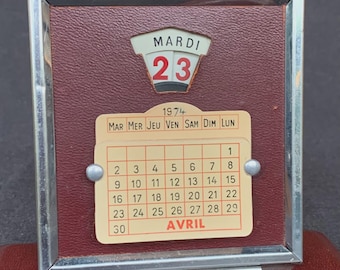 Pode incluir: Um calendário de secretária vintage de 1974, com fundo bordô e moldura prateada. O calendário mostra a data "23" e o mês de abril. O calendário está em francês.