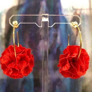 Puede incluir: Un par de pendientes de aro dorados con detalles de flores de tela roja. Los pendientes se muestran en un soporte transparente.
