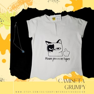 Może przedstawiać: Biała koszulka z czarno-białą grafiką zrzędliwego kota i napisem "Mirame pero no me toques". Widoczny jest również srebrny naszyjnik z niebieskim wisiorkiem. Koszulka ma dekolt w kształcie litery V.