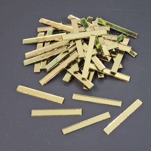 Könnte beinhalten: Ein Haufen goldfarbener, rechteckiger Spiegelkacheln. Die Kacheln sind in einem zufälligen Muster angeordnet und auf einer grauen Oberfläche verstreut.