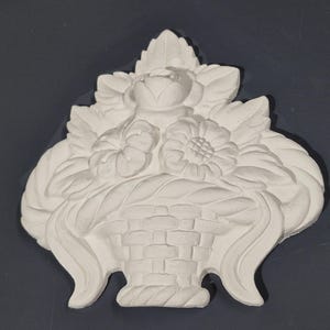 May include: A white plaster wall decoration depicting a basket brimming with flowers and leaves. The design includes a central rose, two sunflowers, and various leaves, all in a detailed, relief style.