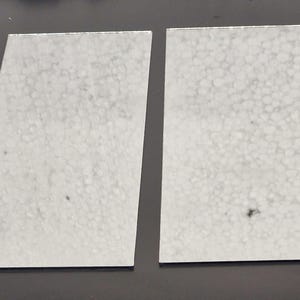 Puede incluir: Dos piezas rectangulares de espuma blanca con una superficie texturizada. La espuma tiene un tono ligeramente gris y pequeñas manchas negras.