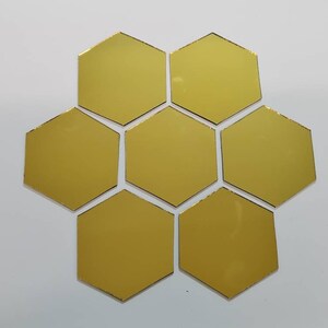 Puede incluir: Siete baldosas doradas en forma de hexágono dispuestas en un patrón circular. Las baldosas tienen una superficie reflectante y un fino borde negro. El fondo es una superficie blanca lisa.