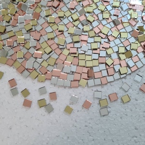 May include: A large pile of small square mirror tiles in gold, silver, and rose gold colours. The tiles are scattered on a white surface.
