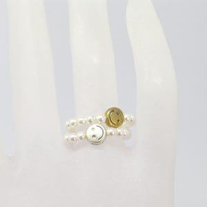 Könnte beinhalten: Zwei Ringe mit Perlendesign und Smiley-Anhängern. Ein Ring hat einen silbernen Smiley-Anhänger, der andere einen goldenen Smiley-Anhänger. Die Ringe befinden sich auf weißem Hintergrund.