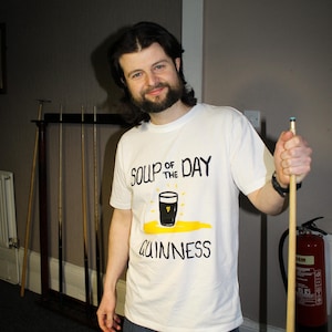 以下が含まれることがあります： 「SOUP OF THE DAY GUINNESS」の文字とギネスビールのパイントのグラフィックがプリントされた白いTシャツ。ビリヤードのキューを持っている。背景には赤い消火器が見える。