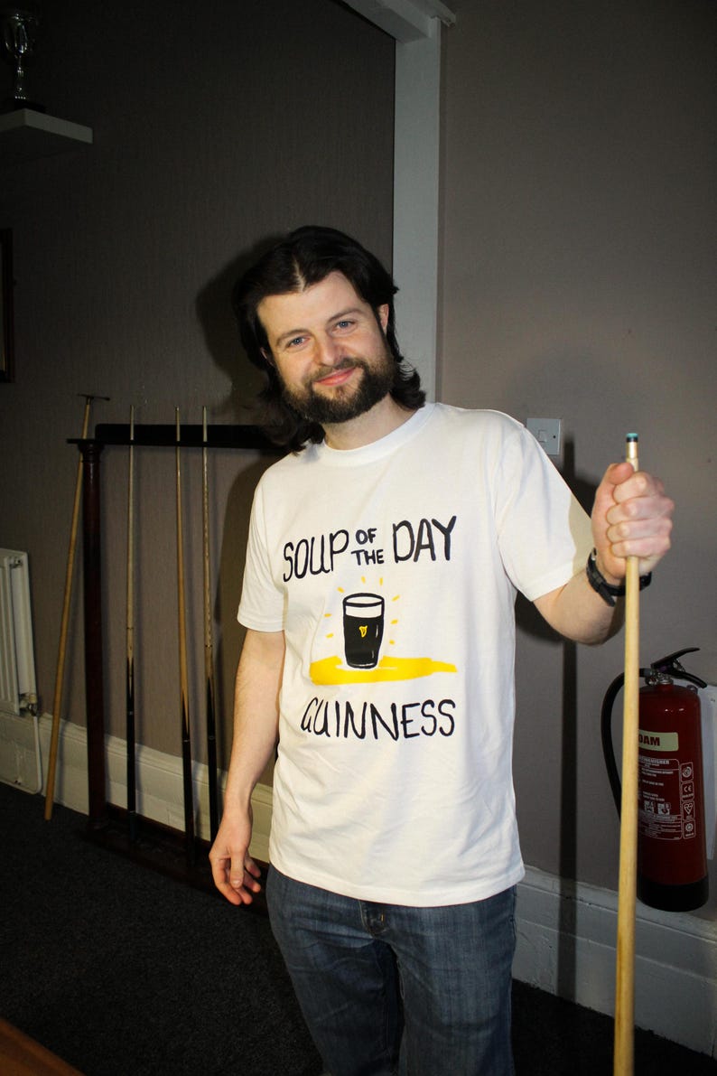 以下が含まれることがあります： 「SOUP OF THE DAY GUINNESS」の文字とギネスビールのパイントのグラフィックがプリントされた白いTシャツ。ビリヤードのキューを持っている。背景には赤い消火器が見える。