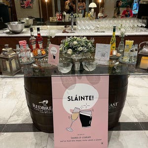 Op de afbeelding: Een bruiloft receptietafel met drankjes, glaswerk en bloemstukken. De tafel wordt ondersteund door houten vaten. Een roze bord met "SL&Aacute;INTE!" en een toast illustratie is zichtbaar, samen met de namen "Laura & Ciaran".