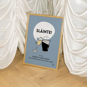 Op de afbeelding: Een ingelijste print met een lichtblauwe achtergrond. Het toont het woord "SL&Aacute;INTE!" boven illustraties van een champagneglas en een pint stout. De print bevat de namen "Terese & Sam Boylan" en de datum "26-06-20". De tekst "we've had the mass, now GRAB a glass!" is ook opgenomen.