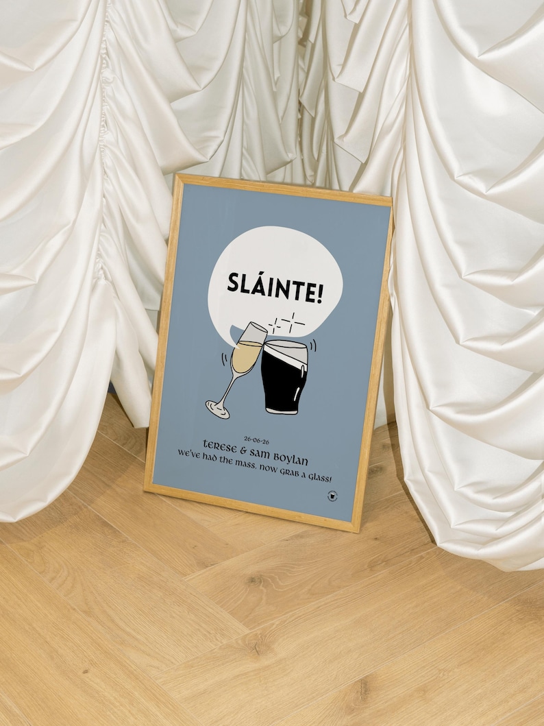 Op de afbeelding: Een ingelijste print met een lichtblauwe achtergrond. Het toont het woord "SL&Aacute;INTE!" boven illustraties van een champagneglas en een pint stout. De print bevat de namen "Terese & Sam Boylan" en de datum "26-06-20". De tekst "we've had the mass, now GRAB a glass!" is ook opgenomen.