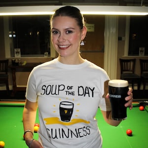 以下が含まれることがあります： 「SOUP OF THE DAY GUINNESS」の文字とギネスビールのパイントのグラフィックがプリントされた白いTシャツ。 ギネスビールのパイントを持っています。 背景には緑色のビリヤード台と赤と黄色のビリヤードボールがあります。