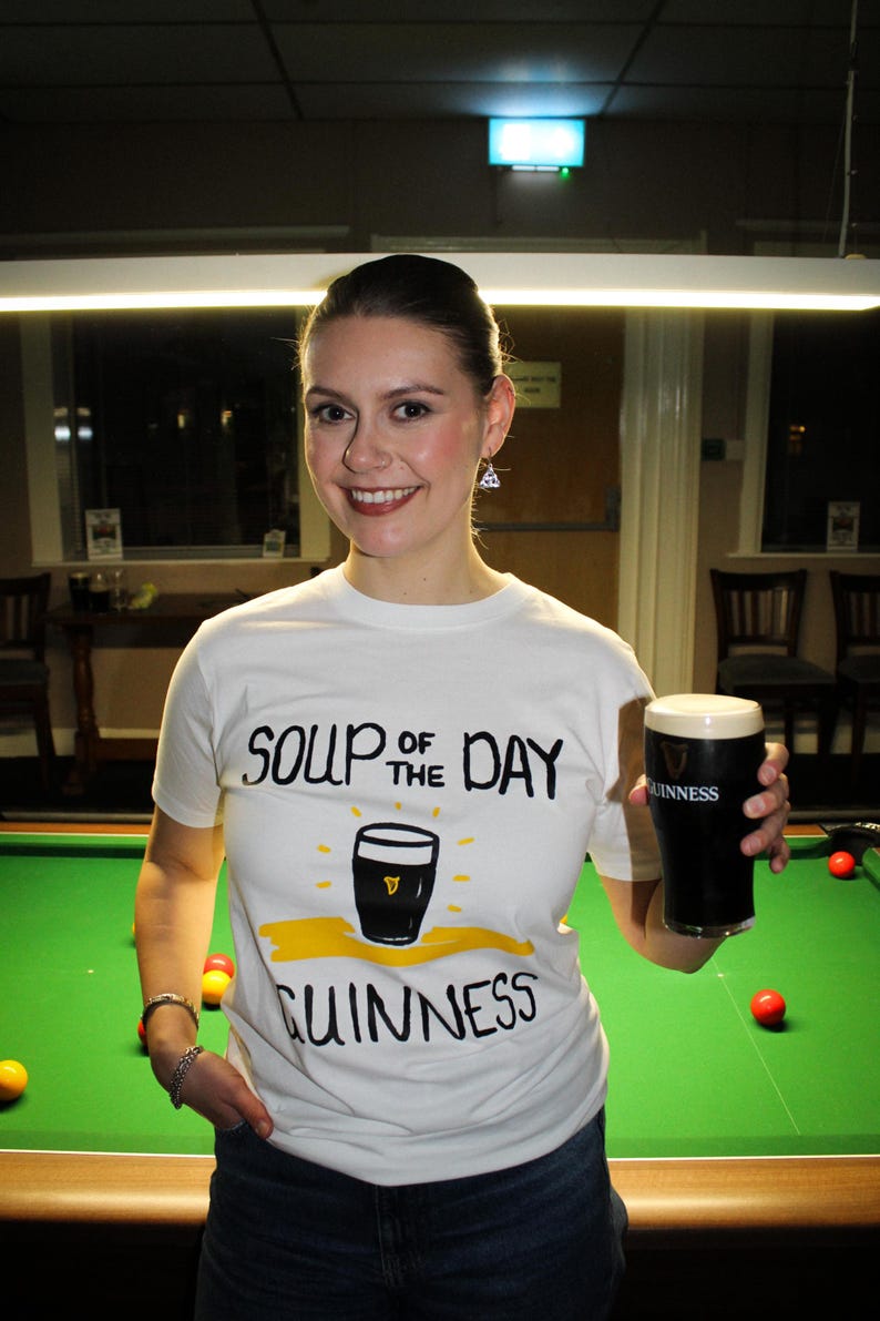 以下が含まれることがあります： 「SOUP OF THE DAY GUINNESS」の文字とギネスビールのパイントのグラフィックがプリントされた白いTシャツ。 ギネスビールのパイントを持っています。 背景には緑色のビリヤード台と赤と黄色のビリヤードボールがあります。
