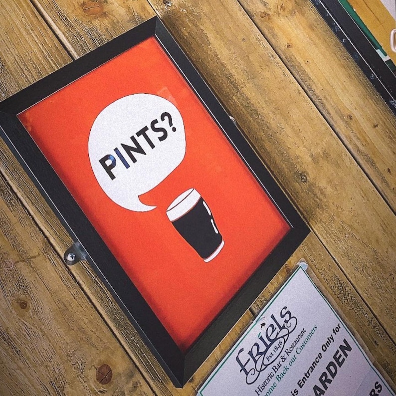 Puede incluir: Cartel enmarcado con fondo rojo y la palabra "PINTS?" en una burbuja blanca. Debajo, un vaso de pinta negro. El cartel est&aacute; en un marco negro y sobre una superficie de madera.