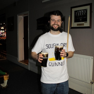 以下が含まれることがあります： ギネスビールを2パイント持っている人。その人は「SOUP of DAY GUINNESS」と書かれた白いTシャツを着ています。ビリヤードのキューも見えます。その人は微笑んでいます。