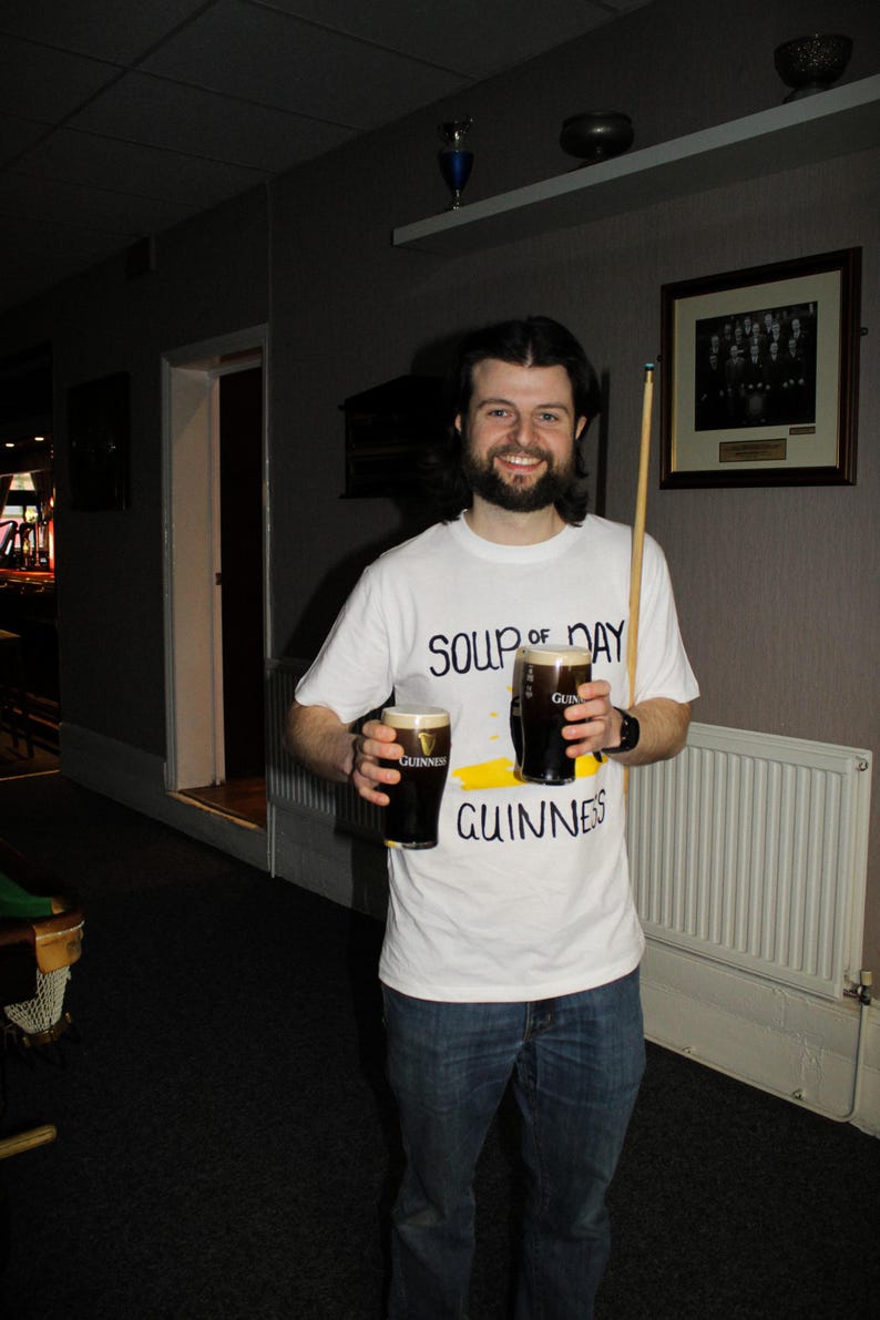 以下が含まれることがあります： ギネスビールを2パイント持っている人。その人は「SOUP of DAY GUINNESS」と書かれた白いTシャツを着ています。ビリヤードのキューも見えます。その人は微笑んでいます。