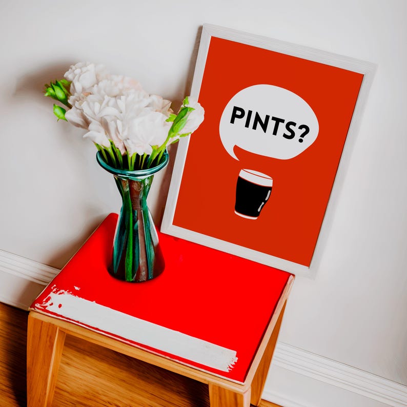 Puede incluir: Un cuadro rojo enmarcado con la palabra "PINTS?" en un bocadillo blanco y una ilustraci&oacute;n de un vaso de pinta negro. El cuadro est&aacute; sobre una mesa roja con un borde blanco, junto a un jarr&oacute;n de cristal con flores blancas.