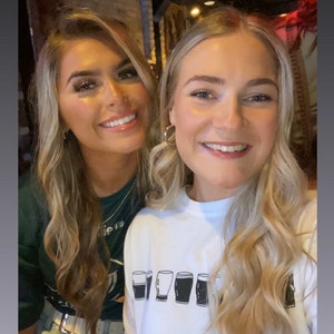 May include: Two women smiling for a photo. The woman on the right is wearing a white shirt with a graphic of five pint glasses. The text "@clodaghcunningham" and three shamrocks are below the graphic.
