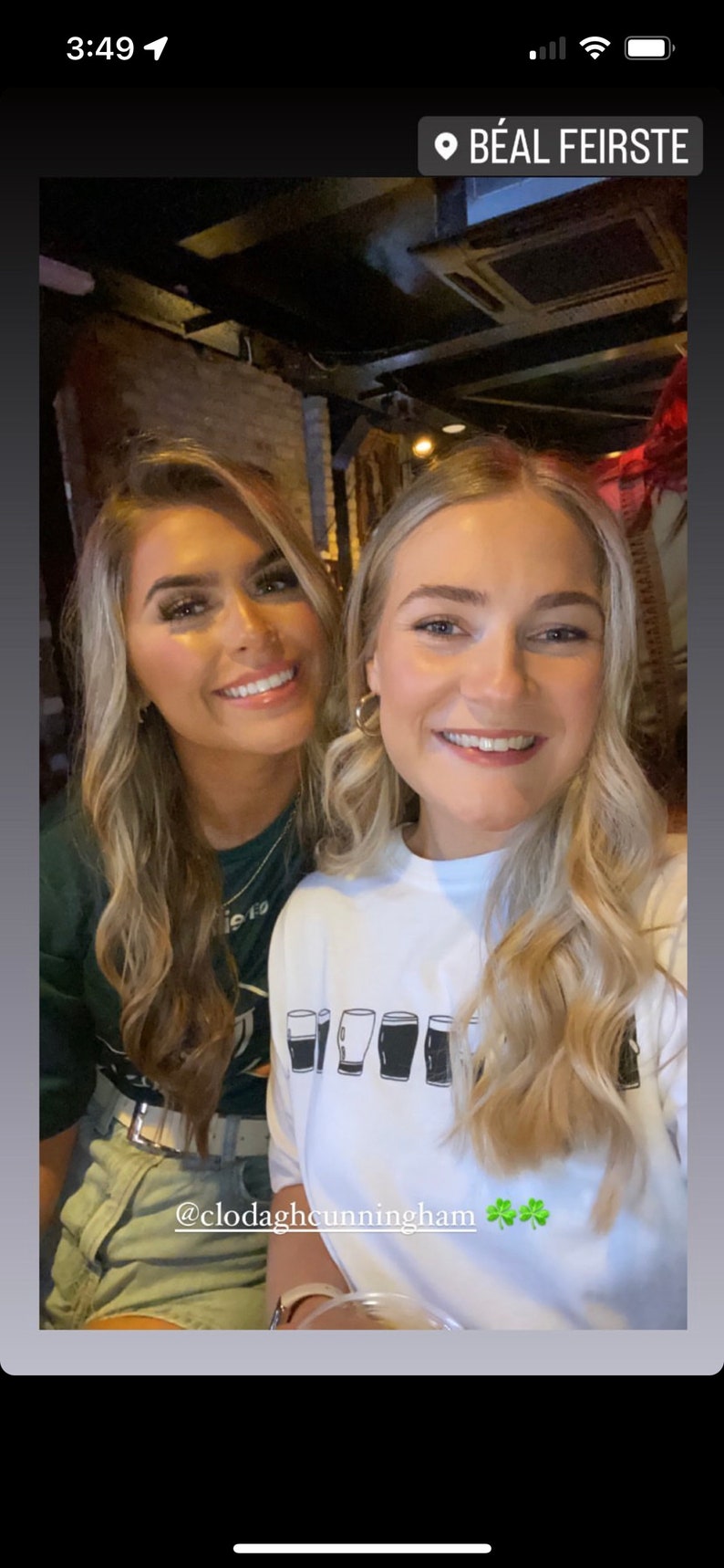 May include: Two women smiling for a photo. The woman on the right is wearing a white shirt with a graphic of five pint glasses. The text "@clodaghcunningham" and three shamrocks are below the graphic.