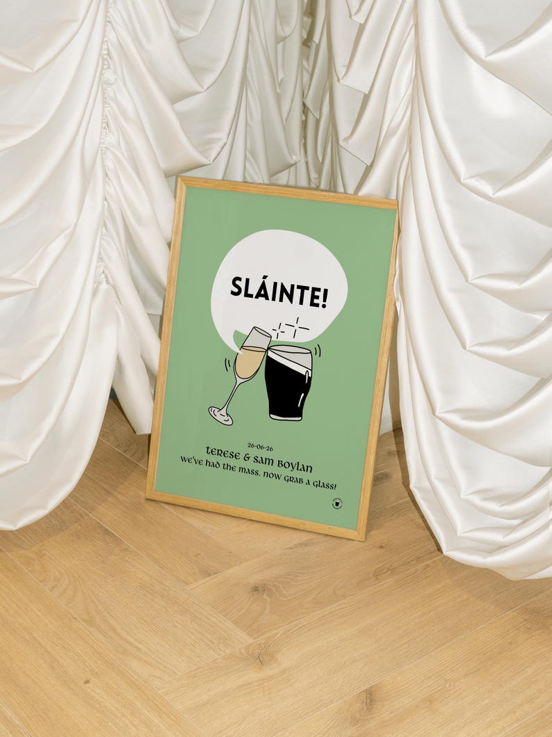 Op de afbeelding: Een ingelijste print met een lichtgroene achtergrond en een houten lijst. De print toont het woord "SL&Aacute;INTE!" in een witte tekstballon, samen met illustraties van een champagneglas en een pint stout. De tekst "terese & sam boylan" en "we've had the mass, now grab a glass!" zijn ook opgenomen.