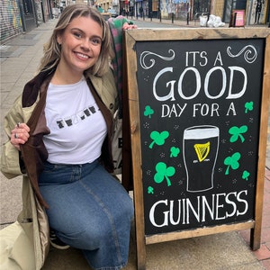 May include: A woman in a brown jacket and blue jeans poses in front of a black chalkboard sign that reads "It's a good day for a Guinness". The sign has a green shamrock design and a glass of Guinness beer.