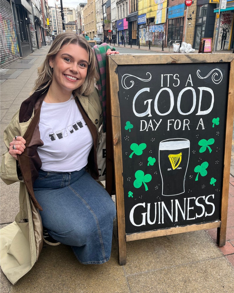 May include: A woman in a brown jacket and blue jeans poses in front of a black chalkboard sign that reads "It's a good day for a Guinness". The sign has a green shamrock design and a glass of Guinness beer.