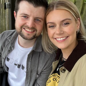 May include: A close-up portrait of a couple smiling at the camera. The man is wearing a gray corduroy jacket over a white t-shirt with a graphic of multiple pint glasses. The woman is wearing a tan jacket with a brown collar over a black t-shirt with a yellow circle graphic.