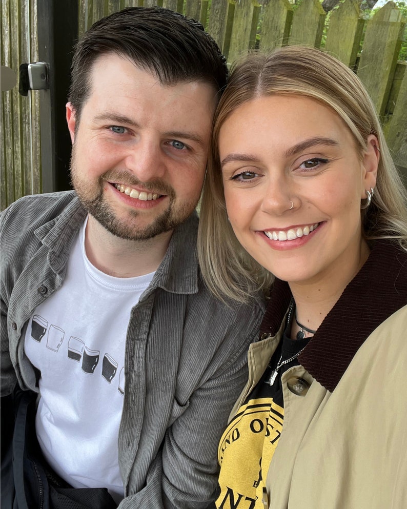 May include: A close-up portrait of a couple smiling at the camera. The man is wearing a gray corduroy jacket over a white t-shirt with a graphic of multiple pint glasses. The woman is wearing a tan jacket with a brown collar over a black t-shirt with a yellow circle graphic.