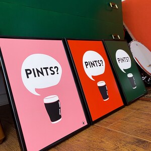 Puede incluir: Tres impresiones art&iacute;sticas enmarcadas con la palabra "PINTS?" en un bocadillo sobre una pinta de cerveza negra. Las impresiones tienen fondos rosa, rojo y verde, con marcos negros. El arte se exhibe sobre un suelo de madera.