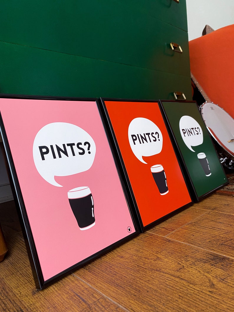 Puede incluir: Tres impresiones art&iacute;sticas enmarcadas con la palabra "PINTS?" en un bocadillo sobre una pinta de cerveza negra. Las impresiones tienen fondos rosa, rojo y verde, con marcos negros. El arte se exhibe sobre un suelo de madera.