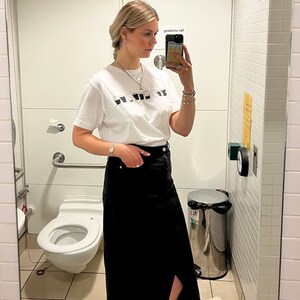 May include: A woman wearing a white t-shirt with a black and white graphic print and a black denim skirt. She is standing in a bathroom and taking a selfie.
