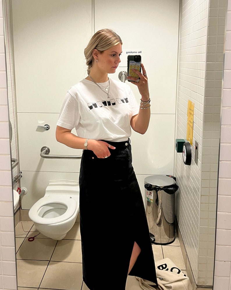 May include: A woman wearing a white t-shirt with a black and white graphic print and a black denim skirt. She is standing in a bathroom and taking a selfie.