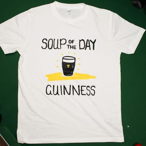 以下が含まれることがあります： 黒と黄色のグラフィックが施された白いTシャツ。グラフィックは、上部に「SOUP OF THE DAY」、下部に「GUINNESS」と書かれたギネスビールのパイントグラスが特徴です。シャツは赤いボールのある緑色の表面にあります。