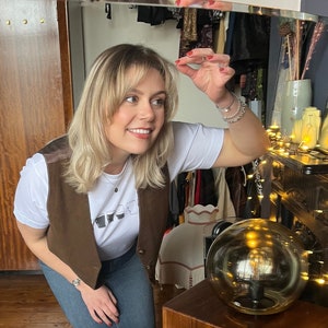 May include: A woman wearing a brown vest over a white t-shirt with a graphic of glasses. She is wearing blue jeans and has blonde hair with bangs. She is standing in front of a mirror and looking at herself. There is a light fixture with a round glass shade in the background.
