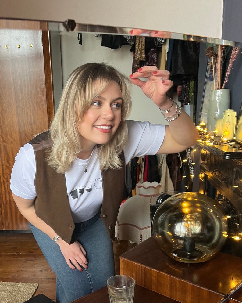 May include: A woman wearing a brown vest over a white t-shirt with a graphic of glasses. She is wearing blue jeans and has blonde hair with bangs. She is standing in front of a mirror and looking at herself. There is a light fixture with a round glass shade in the background.