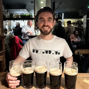 May include: A man wearing a white t-shirt with a row of black Guinness pint glasses printed on the front, sits at a bar with four full glasses of Guinness in front of him.