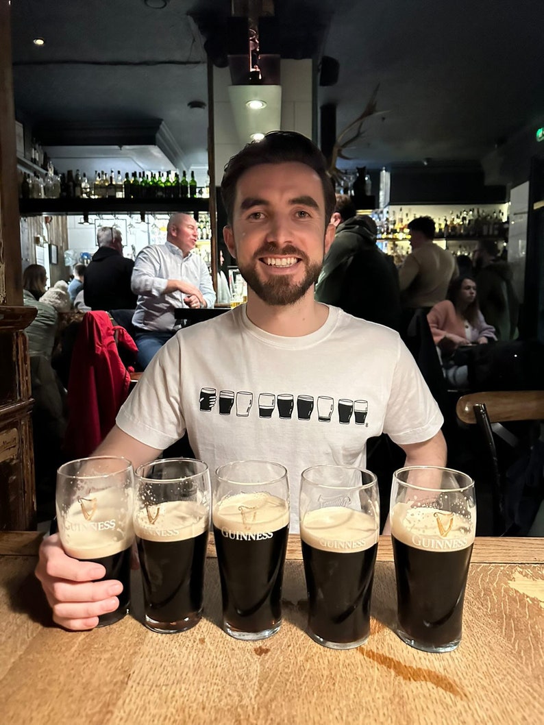 May include: A man wearing a white t-shirt with a row of black Guinness pint glasses printed on the front, sits at a bar with four full glasses of Guinness in front of him.