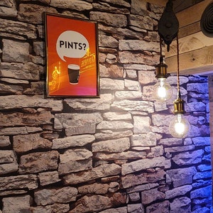 Puede incluir: Un p&oacute;ster enmarcado con el texto "PINTS?" encima de un gr&aacute;fico de una pinta de cerveza negra. El p&oacute;ster cuelga de una pared de piedra, con dos l&aacute;mparas colgantes con bombillas Edison colgando del techo. El techo es de madera.