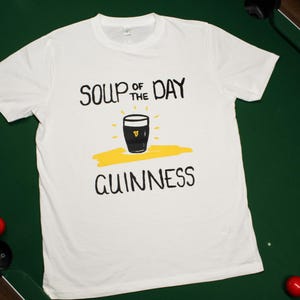 以下が含まれることがあります： 黒い文字で「SOUP OF THE DAY GUINNESS」と書かれた白いTシャツ。黄色いアクセントのギネスビールのパイントのグラフィックが特徴です。ビリヤードのボールがある緑色の表面にTシャツが置かれています。