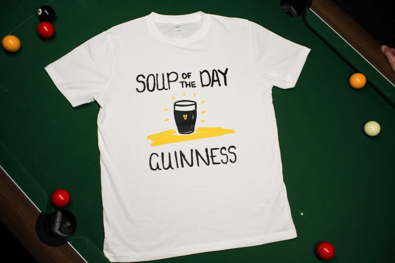 以下が含まれることがあります： 黒い文字で「SOUP OF THE DAY GUINNESS」と書かれた白いTシャツ。黄色いアクセントのギネスビールのパイントのグラフィックが特徴です。ビリヤードのボールがある緑色の表面にTシャツが置かれています。