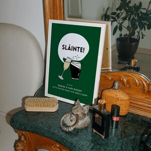 Op de afbeelding: Een ingelijste print met een groene achtergrond en het woord "SL&Aacute;INTE!" in het wit. De print bevat illustraties van een champagneglas en een pint stout. De tekst hieronder luidt "terese & sam boylan" en "we've had the mass, now grab a glass!"