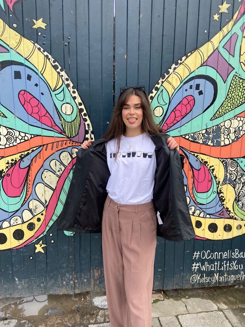 May include: A woman wearing a white t-shirt with a graphic of glasses, a black jacket, and pink pants stands in front of a colorful mural with a butterfly design. The mural has the text #OConnellsBar #WhatLiftsYou @KelseyMontagueArt.