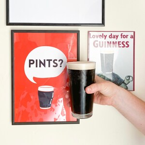 Puede incluir: Una pinta de cerveza negra oscura con una cabeza cremosa se sostiene frente a dos impresiones enmarcadas. Una impresi&oacute;n es roja con la palabra "PINTS?" en una burbuja blanca. La otra impresi&oacute;n dice "Lovely day for a GUINNESS".