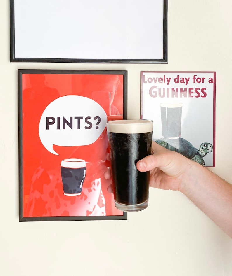 Puede incluir: Una pinta de cerveza negra oscura con una cabeza cremosa se sostiene frente a dos impresiones enmarcadas. Una impresi&oacute;n es roja con la palabra "PINTS?" en una burbuja blanca. La otra impresi&oacute;n dice "Lovely day for a GUINNESS".