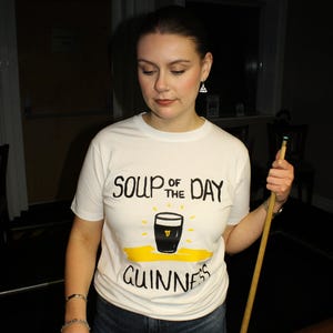 以下が含まれることがあります： 「SOUP OF THE DAY」の文字とギネスビールのパイントのグラフィック、そして「GUINNESS」の文字がプリントされた白いTシャツ。人物はギネスビールのパイントとビリヤードのキューを持っています。