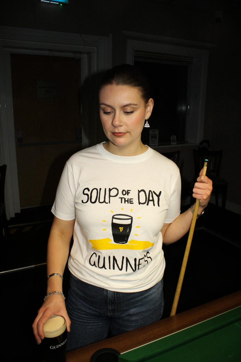 以下が含まれることがあります： 「SOUP OF THE DAY」の文字とギネスビールのパイントのグラフィック、そして「GUINNESS」の文字がプリントされた白いTシャツ。人物はギネスビールのパイントとビリヤードのキューを持っています。
